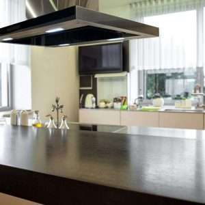 Closeup of black wooden tabletop of kitchen island with cooktop and cooker hood on blurred background of light spacious kitchen interior
